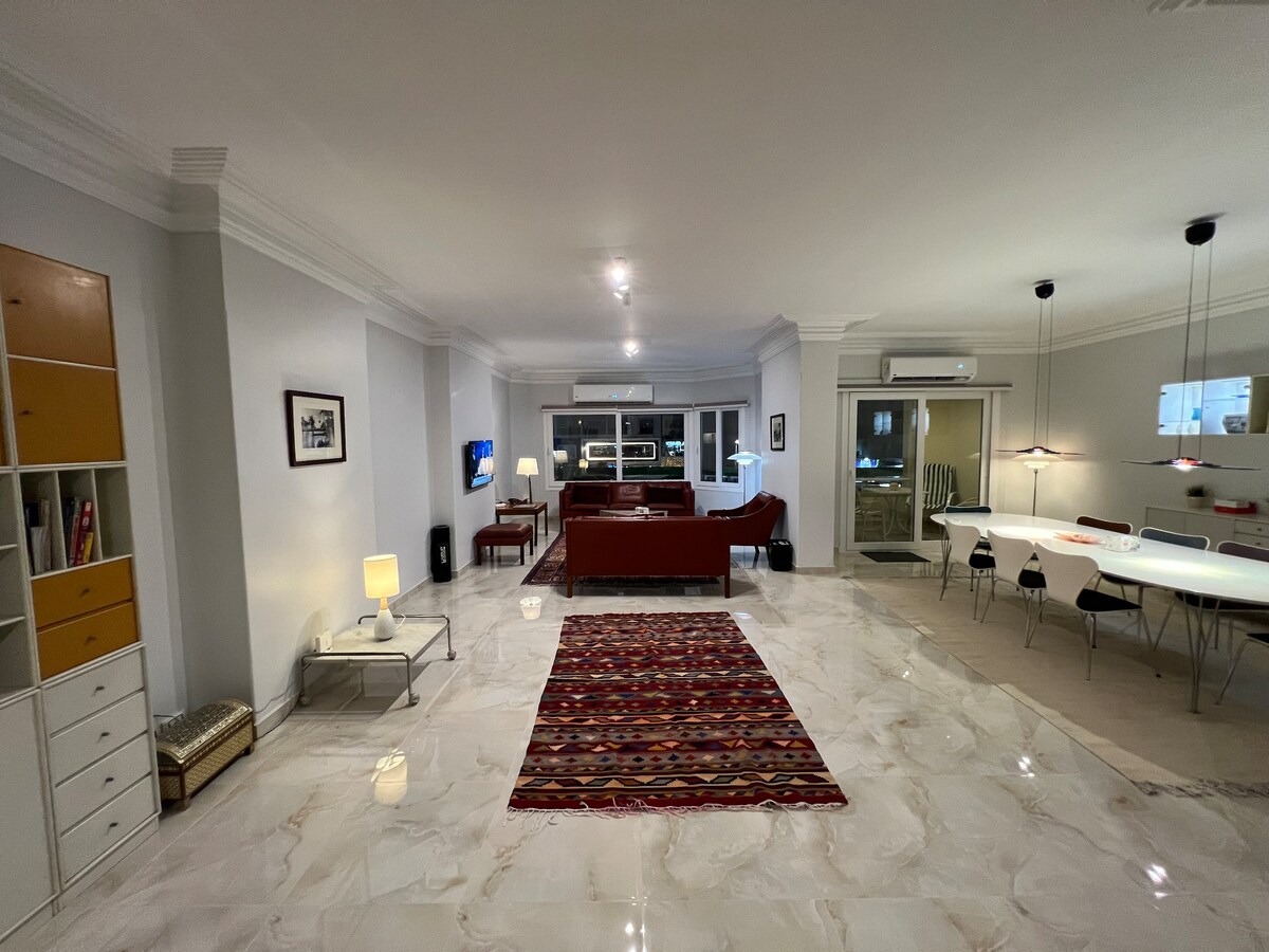 A spacious living area features polished marble floors and elegant Danish design furniture. A red sofa in the center is complemented by a colorful area rug. The dining area consists of a modern table and pendant lights, with large windows allowing natural light to fill the room.