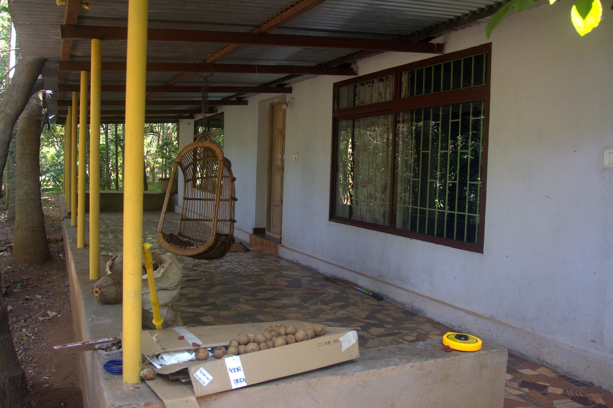 A covered porch area features a traditional hanging swing made of woven material. The floor is tiled with a pattern, and several potted plants are positioned nearby. A large window with green bars offers views into the interior space.