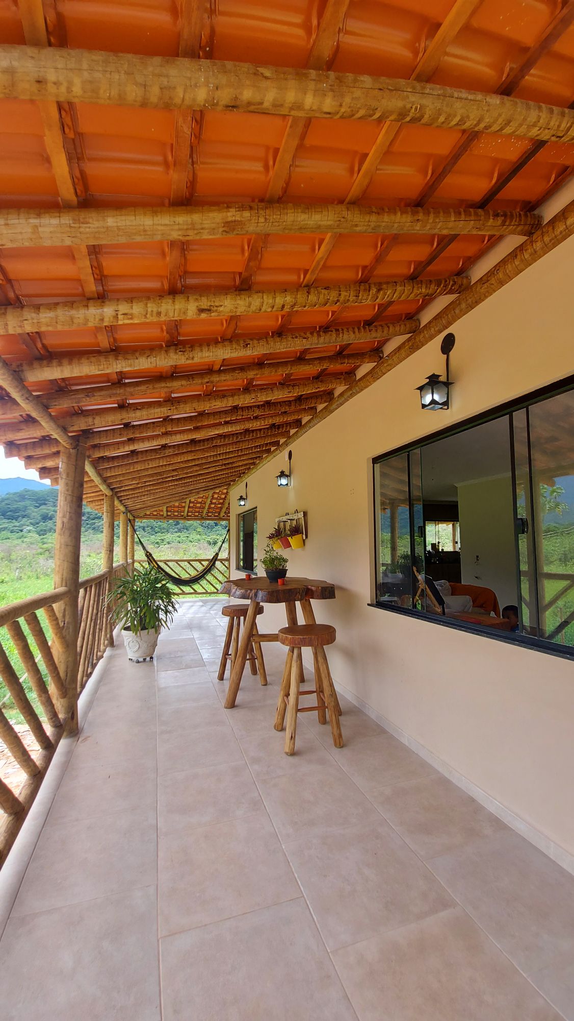 A spacious porch features a wooden railing and a terracotta roof. Two tall stools made of wood are positioned next to a small table. A hammock can be seen hanging to the side, with views of the surrounding lush greenery visible through the large window.