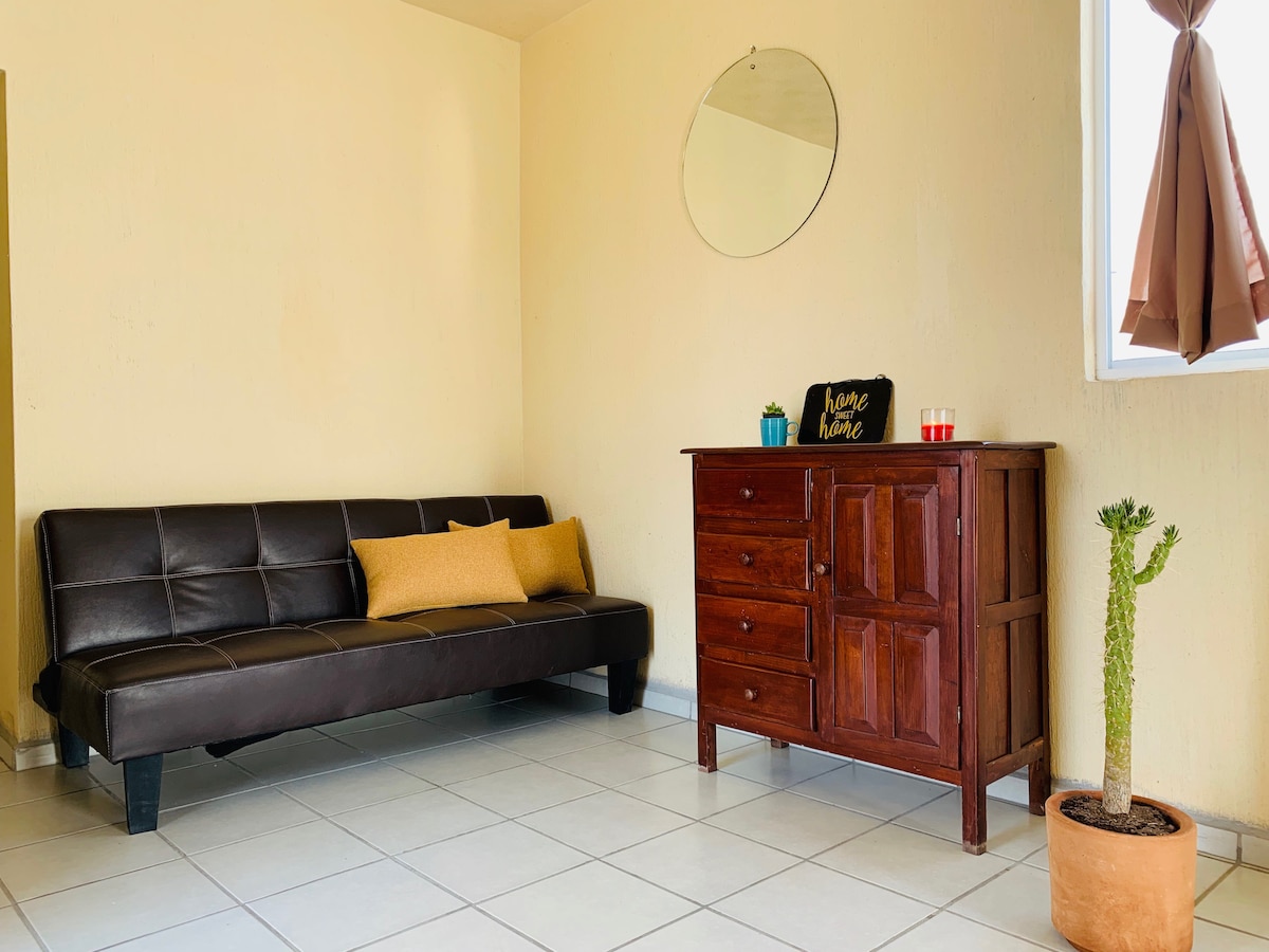 A light and airy living space features a brown sofa bed with a yellow cushion, accompanied by a wooden dresser. A round mirror reflects natural light, while a potted cactus adds greenery. A window with brown curtains allows for varied light control.