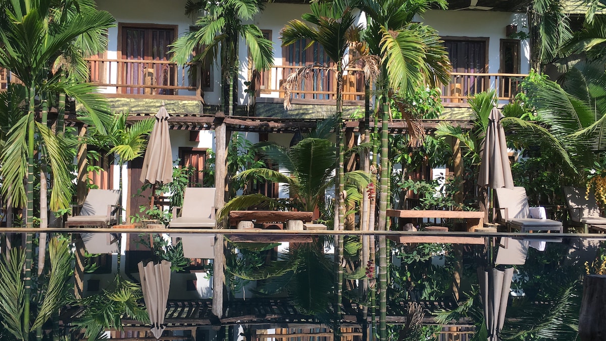 A tranquil pool reflects the surrounding tropical garden and shaded lounge chairs. The multi-level building is framed by lush greenery, with wooden balconies offering views of the serene area. Palms gently sway, enhancing the peaceful ambiance of the setting.