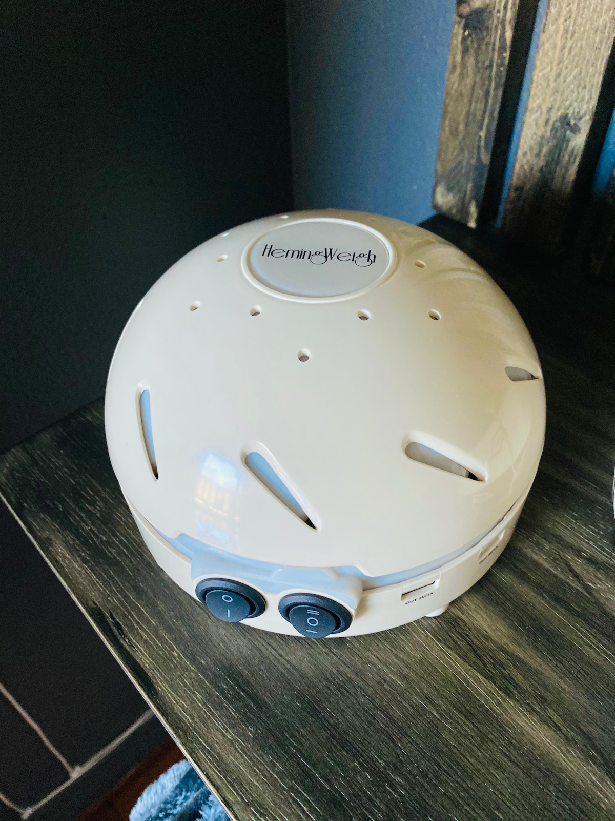 A small, cream-colored white noise machine rests on a dark wooden surface, with a rounded design and a smooth finish. Two buttons are visible on the front, and several small holes are present on the top for sound emission.