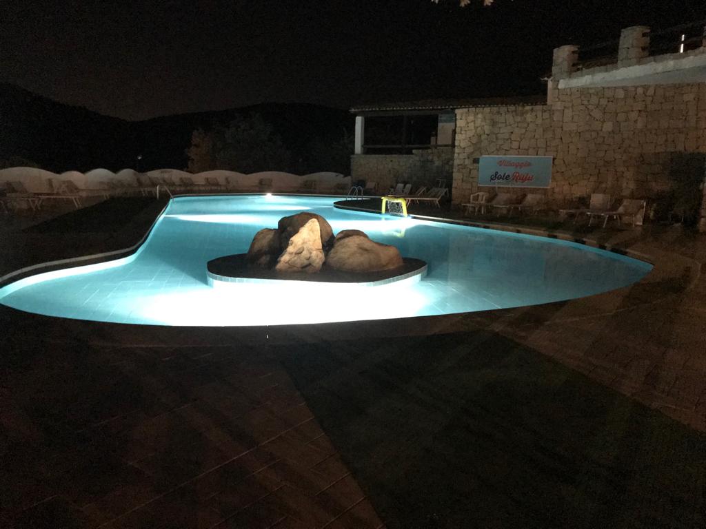A tranquil pool area is illuminated at night, featuring smooth blue water and natural stone accents at its center. Surrounding lounge chairs are positioned around the pool, with soft lighting enhancing the serene ambiance. The backdrop includes a stone wall and hints of surrounding hills.