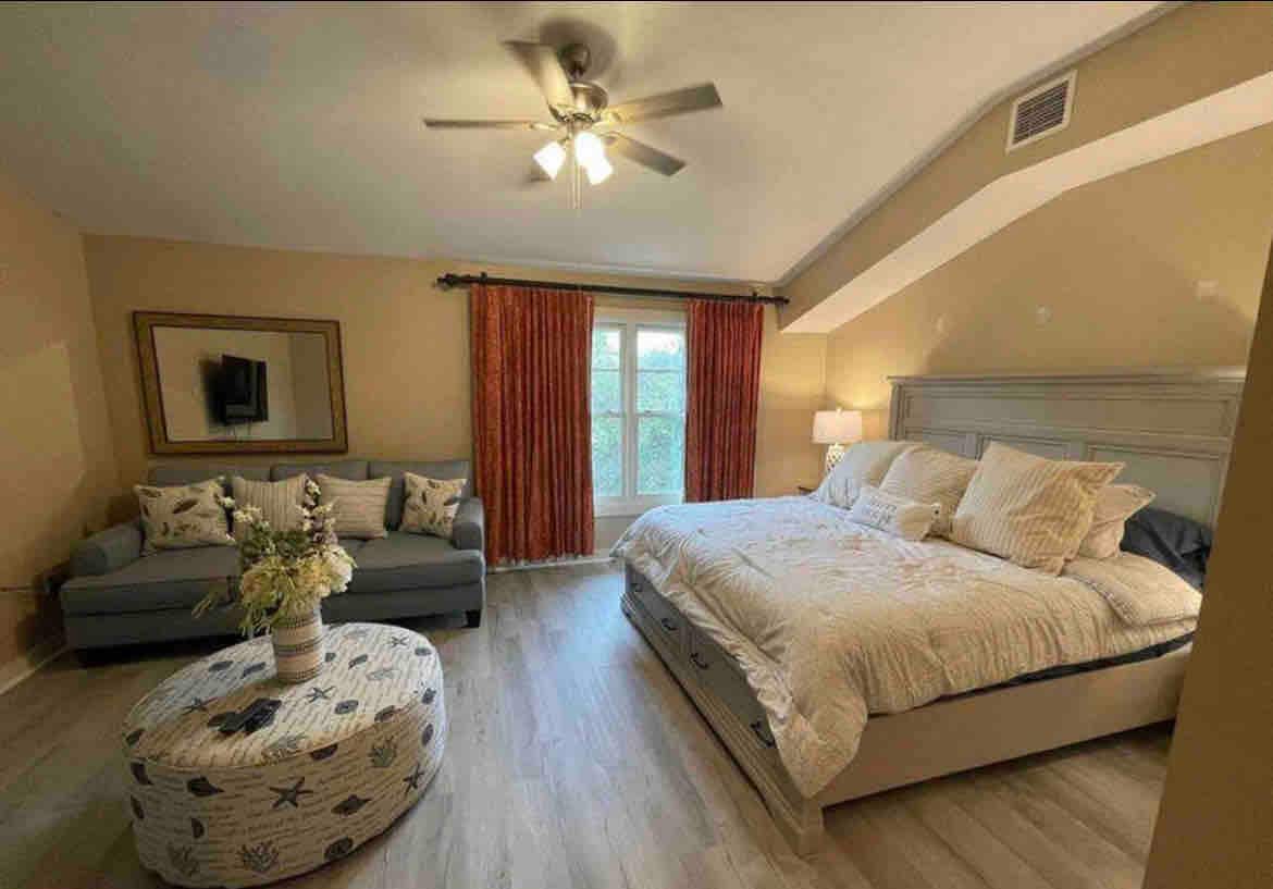 A spacious bedroom features a large bed adorned with neutral linens and decorative pillows. A plush loveseat is situated to one side, complemented by a round ottoman in front. Natural light enters through large windows, framed by soft red curtains, while a ceiling fan adds a cooling touch.