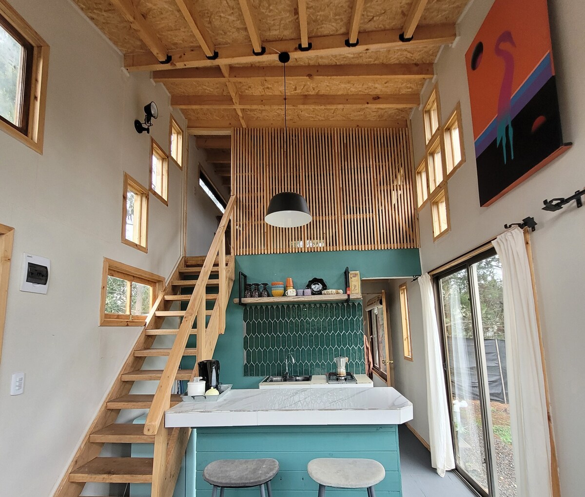 An interior view showcases a modern kitchen area with a large island and two stools. The wooden staircase leads to a loft area, and large windows allow natural light to fill the space. A colorful artwork adorns the wall, adding visual interest.