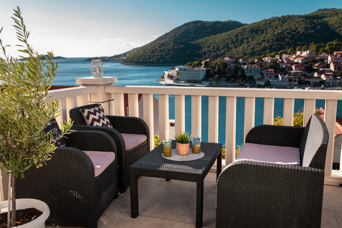A terrace is shown featuring comfortable outdoor seating in black wicker. A small table with decorative plants and refreshments is positioned in the center. The view captures the coastline and distant hills, with the shimmering Adriatic Sea visible in the background.