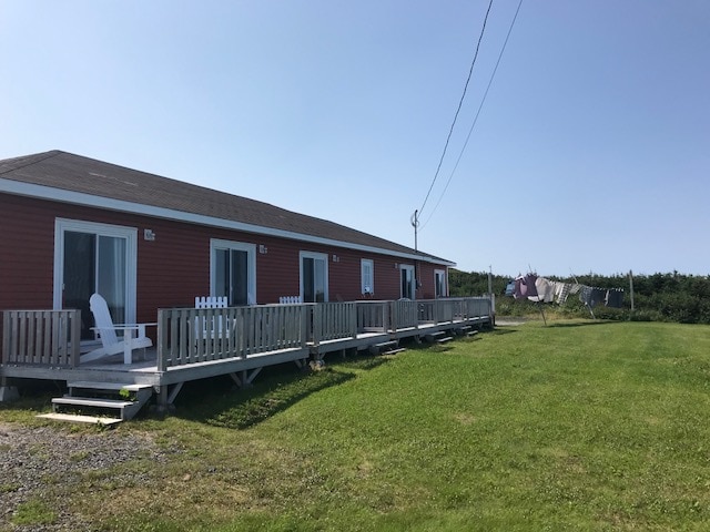 A row of charming red cottages is depicted, each featuring a private deck with white outdoor chairs. Stretching green grass leads to a visible clothesline with hanging laundry. The clear blue sky adds a bright backdrop to the scene.