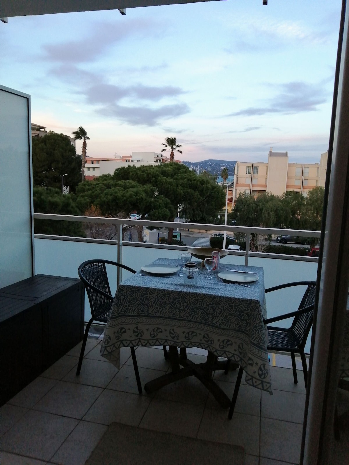 An outdoor balcony features a small dining table set for two, complete with plates and glasses. Surrounding greenery and distant buildings create a calming backdrop, while the sky exhibits soft hues of blue and pink as twilight approaches.