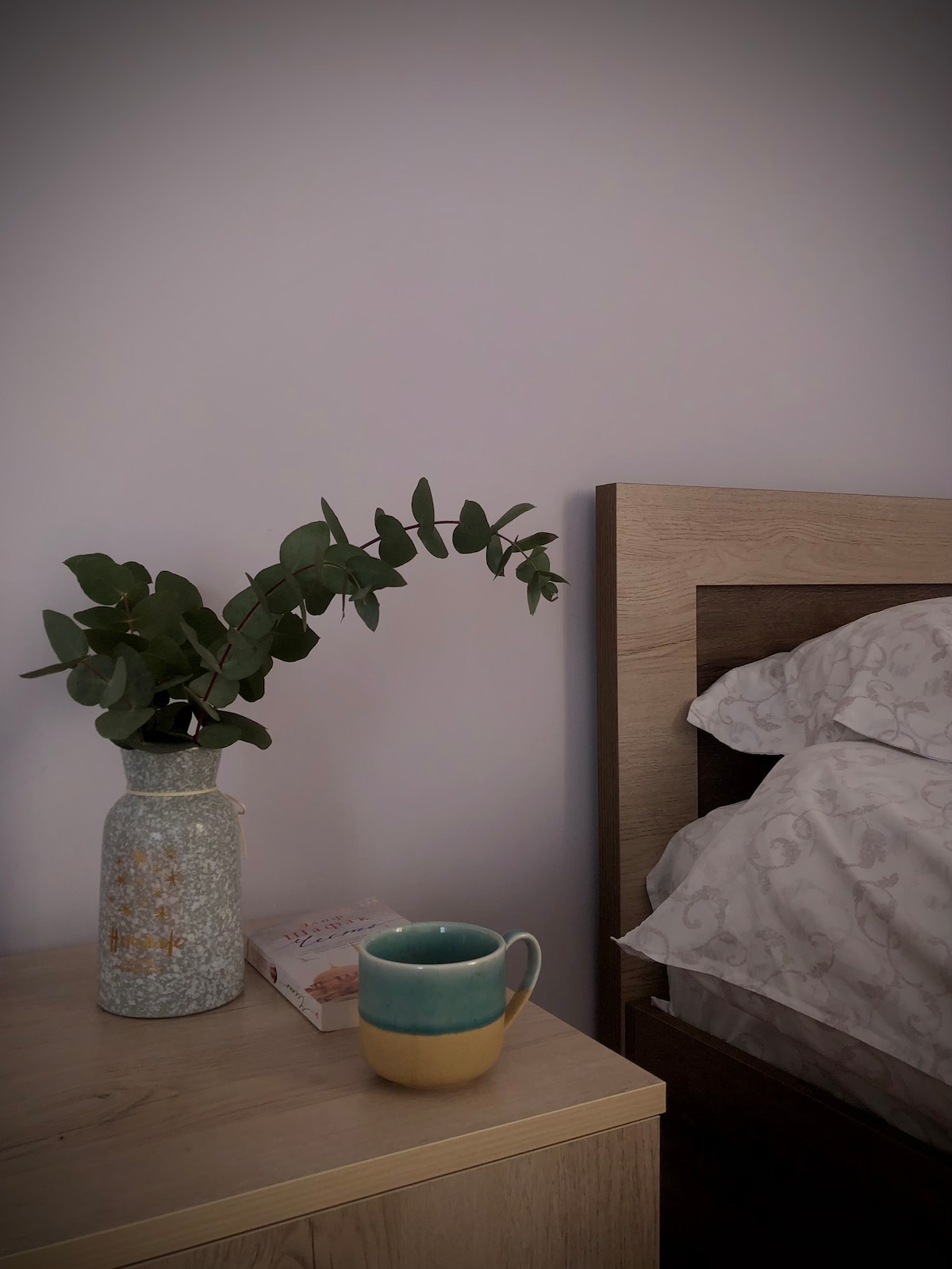 A bedside table is adorned with a textured vase holding fresh greenery. A two-toned mug rests nearby, complementing the serene atmosphere. The bed is dressed in soft, patterned linens. The background features a subtly colored wall, enhancing the calming environment.