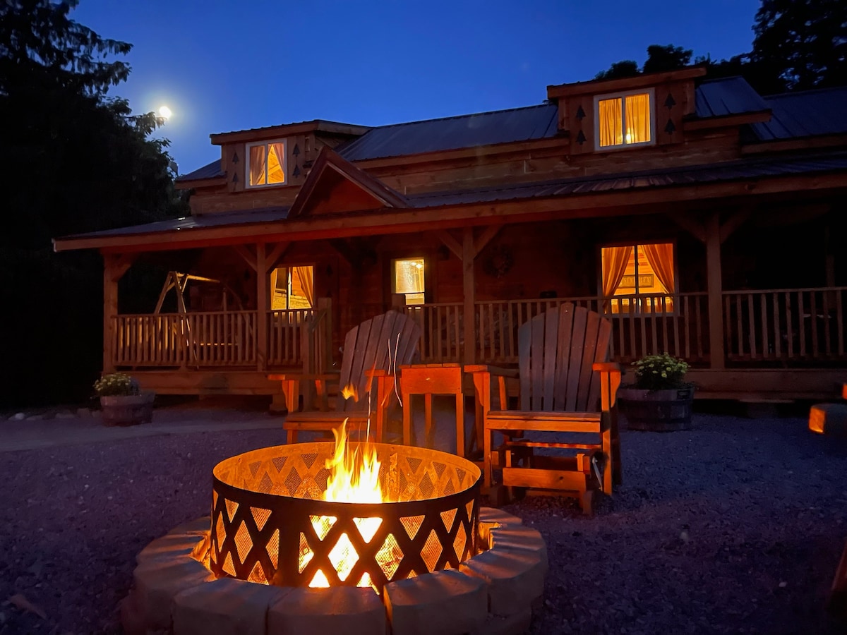 A rustic cabin exterior is illuminated under the night sky, with warm light glowing from the windows. A fire pit is centrally located, surrounded by two wooden Adirondack chairs. The porch features a welcoming swing and potted plants for added charm.