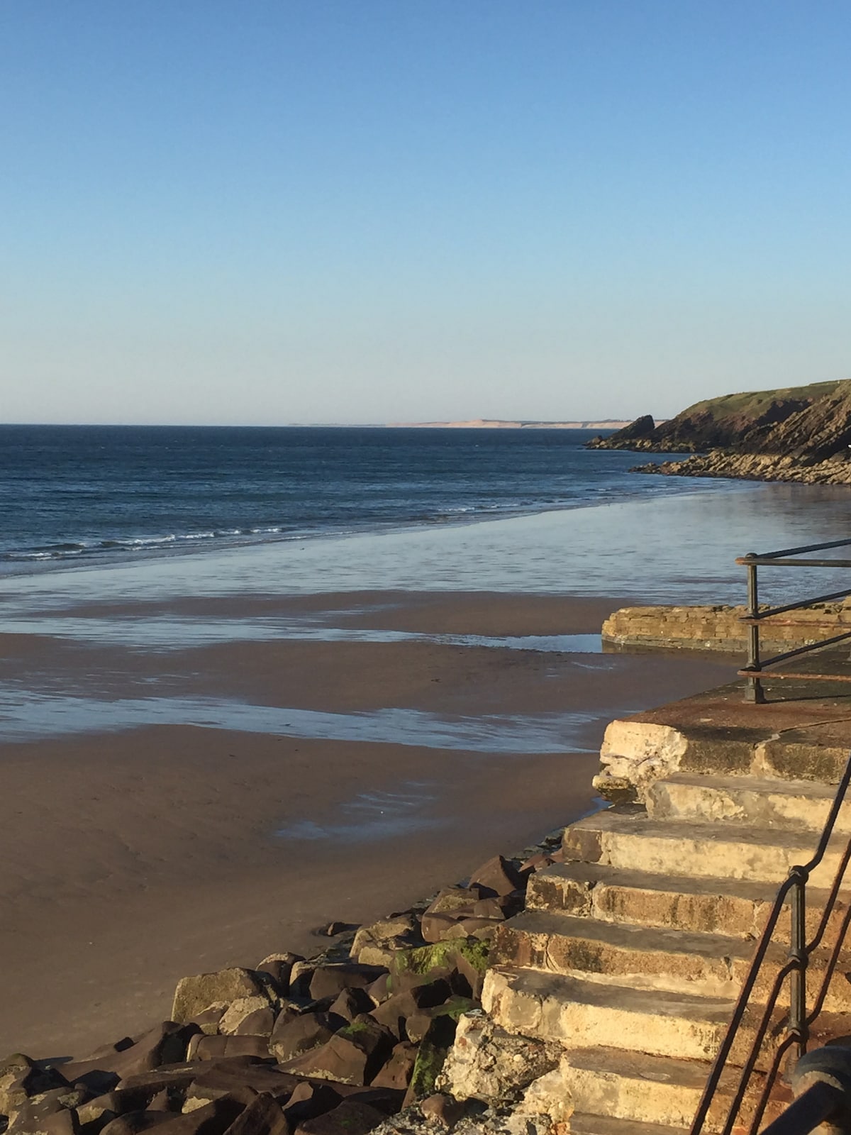 A calm beach scene captures gentle waves lapping against the sandy shoreline. The water reflects a soft blue hue, while rocky cliffs frame the background. A flight of stone steps leads down to the beach, inviting a closer look at the serene surroundings.