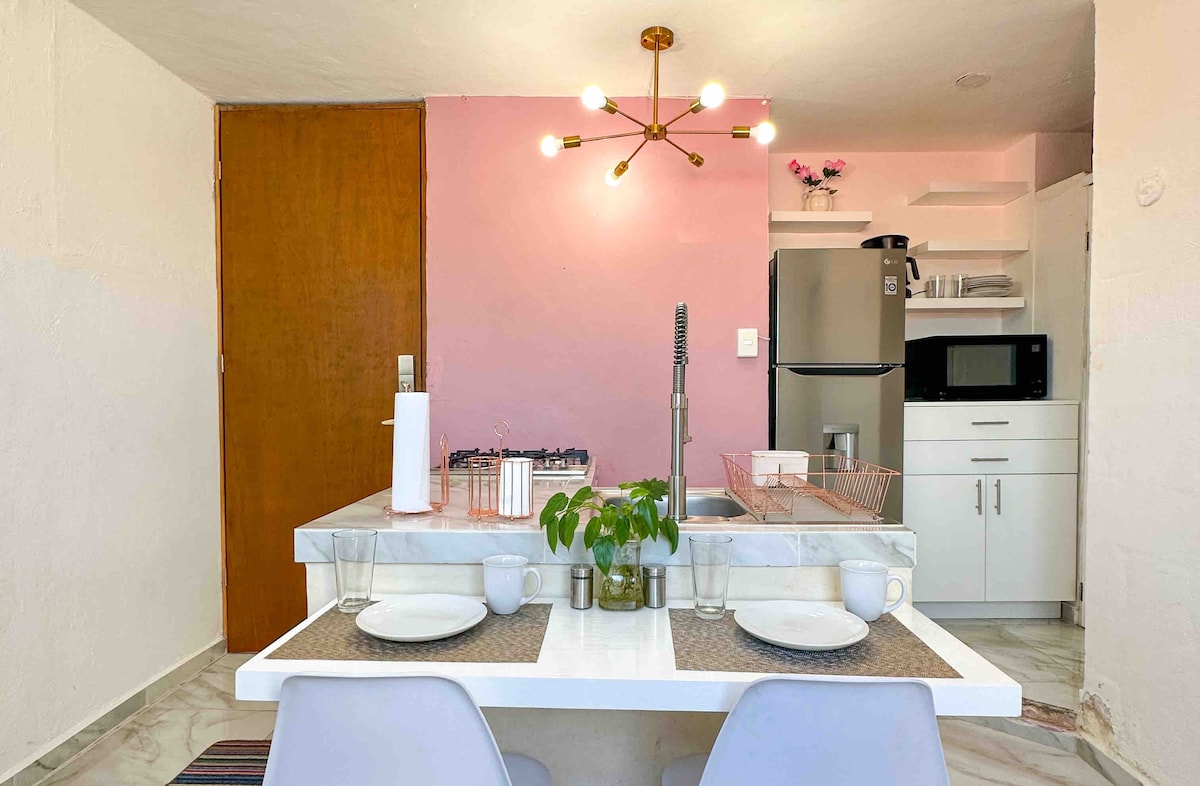 A modern kitchen is presented with a marble dining table set for two. Behind, a pink wall adds a subtle touch, complemented by a stylish light fixture above. Kitchen appliances such as a refrigerator and microwave are neatly arranged, along with a gas stove and sink.