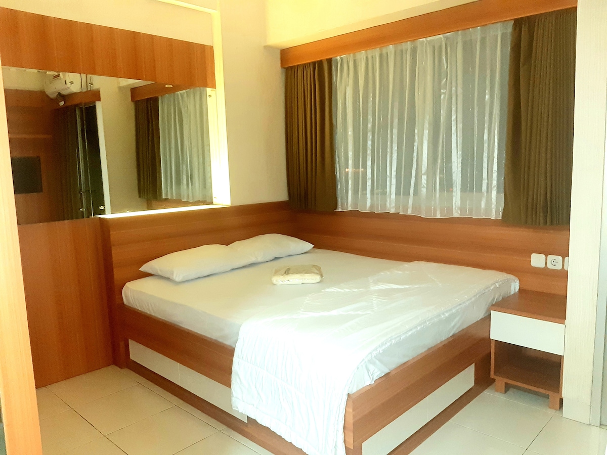 A spacious bedroom features wooden paneling and a large window covered with sheer curtains, allowing natural light to enter. The bed is neatly arranged with white linens, complemented by a towel placed on top. Reflections in the mirrors add depth to the room.