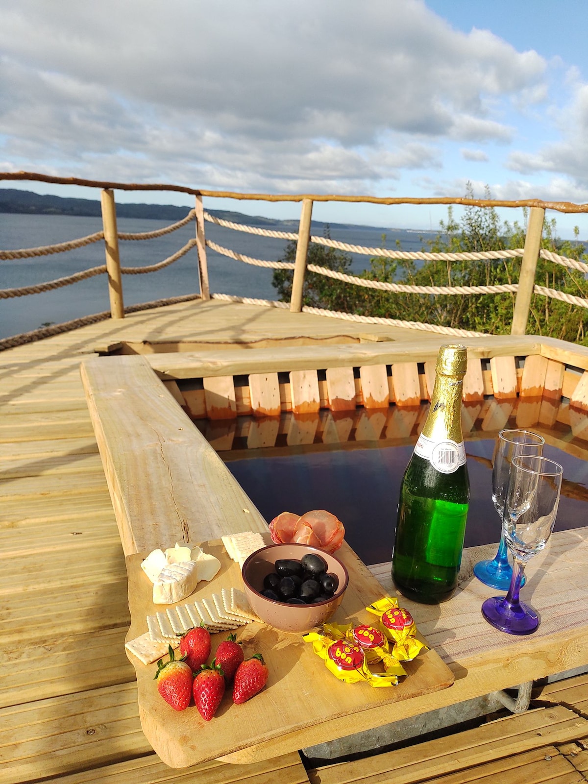 A wooden terrace features a cozy hot tub filled with water, surrounded by a rustic railing. A bottle of sparkling wine stands beside two glasses. Fresh strawberries, olives, and assorted sweets are arranged on a plate, with a tranquil view of the water visible in the background.