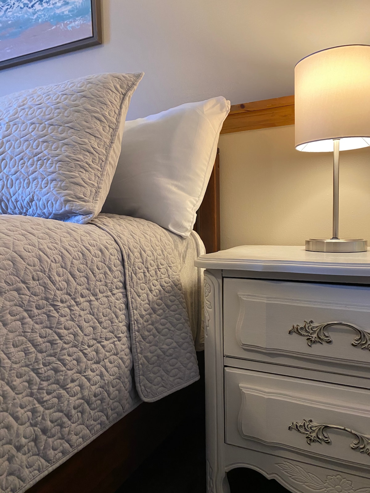 A cozy corner of a bedroom features a spacious bed with a light-colored quilt and plush pillows. A nightstand with decorative molding sits beside the bed, complemented by a softly glowing lamp. Warm light enhances the serene atmosphere of the space.