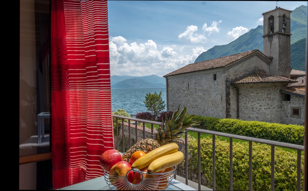 A fruit basket filled with various fresh fruits sits on a table, with a view of a historic stone building and the shimmering waters of Lake Iseo in the background. Mounts rise majestically in the distance under a partly cloudy sky, creating a serene setting.