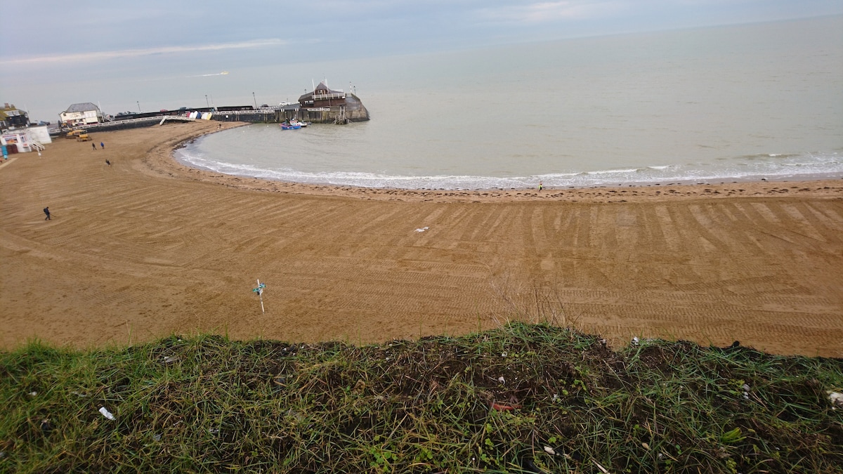 A sandy beach is visible, gently sloping towards the calm sea. Beach huts and a pier are seen in the distance, with a few individuals strolling along the shore. The scene is framed by lush green grass on a gentle incline that offers a view of the beach below.