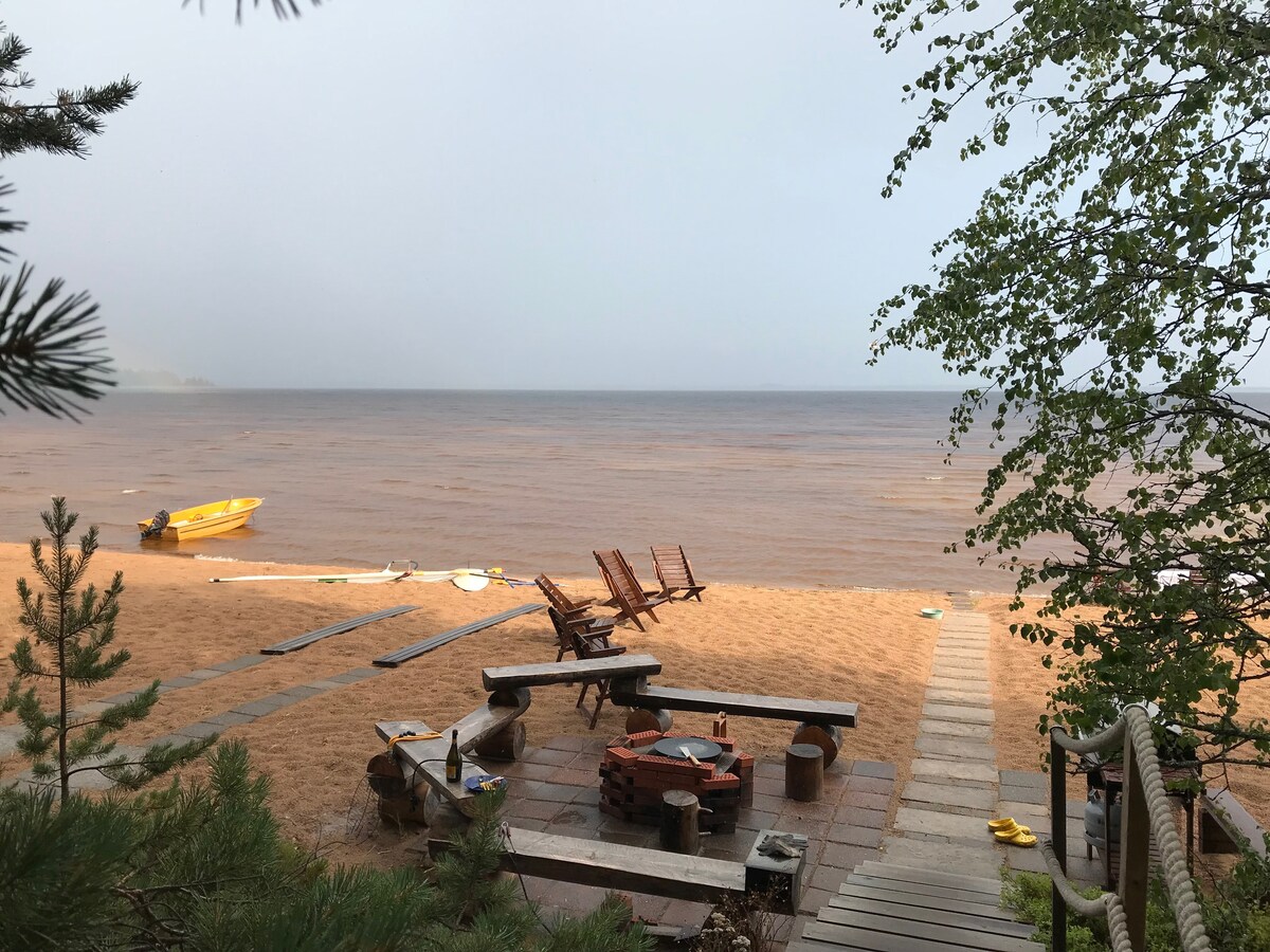 A sandy beach area extends towards a calm water surface, with a clear view of the shoreline. Wooden lounge chairs are arranged near a fire pit, while a yellow boat is anchored nearby. A wooden pathway leads down to the beach, surrounded by greenery.
