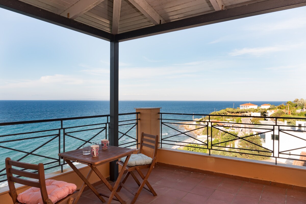 A sea-facing balcony is presented with a wooden table and two chairs. The expansive view of the ocean is visible, with hints of coastal land in the background. The sky features a soft gradient of blue, indicating a clear day.