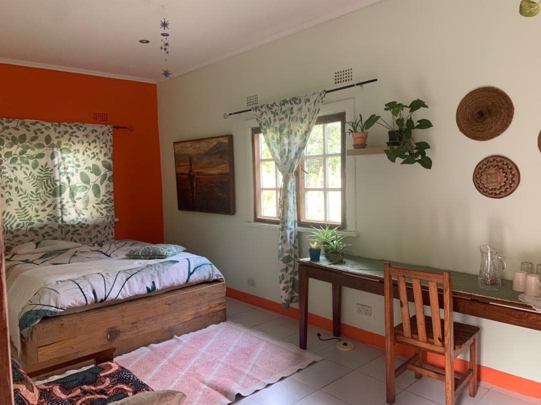 A self-contained room features a wooden bed with a botanical-patterned duvet. A small desk and chair are positioned against the wall. Natural light filters through a window adorned with curtains, while potted plants and wall decor add a touch of greenery to the space.