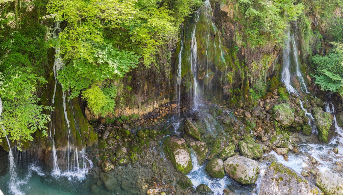 A serene landscape features multiple cascading waterfalls surrounded by lush green foliage. Smooth stones are scattered across the riverbed, and the water creates gentle ripples as it flows over the rocks, offering a refreshing natural atmosphere.