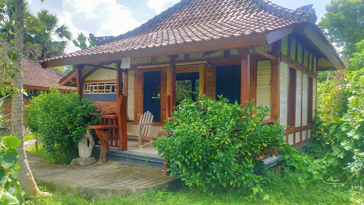 The wooden cottage features a traditional thatched roof and is surrounded by lush greenery. A small wooden deck is present at the entrance, providing space for outdoor seating. Natural light is welcomed through large front doors, enhancing the connection with the peaceful garden.