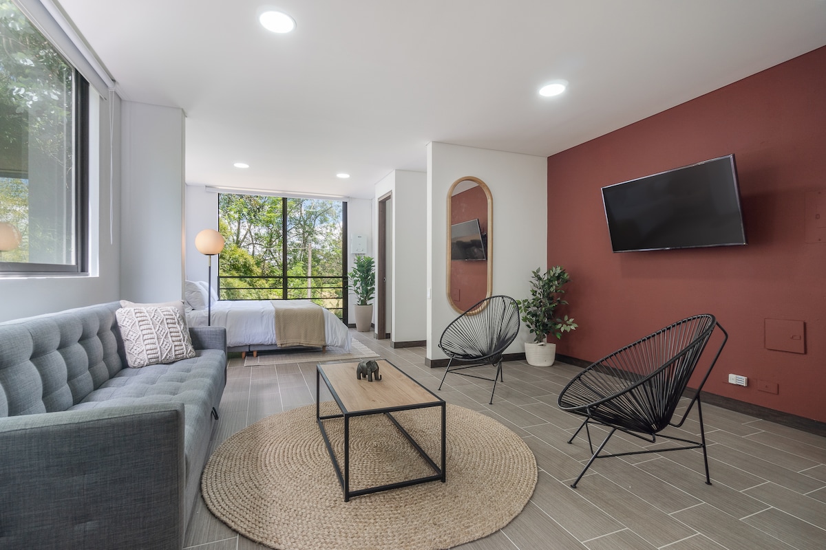 A modern living space features large windows allowing natural light to fill the room. A cozy sofa and two accent chairs surround a central coffee table, enhancing the inviting atmosphere. A TV is mounted on the wall, with tasteful decorative elements positioned throughout the space.