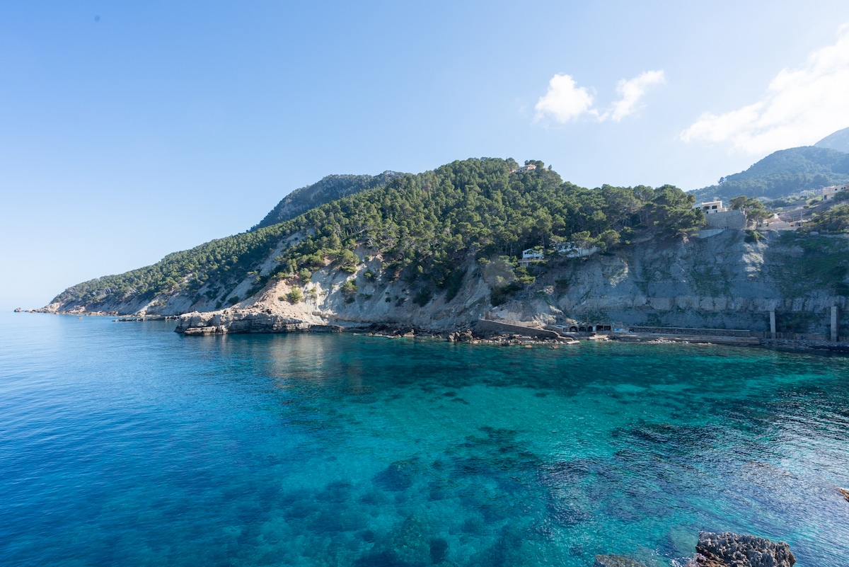 A serene coastal view showcases calm turquoise waters against a steep, rugged cliffside. Lush greenery clings to the mountainside, while a clear blue sky extends overhead. The tranquil scene invites exploration of the natural surroundings.