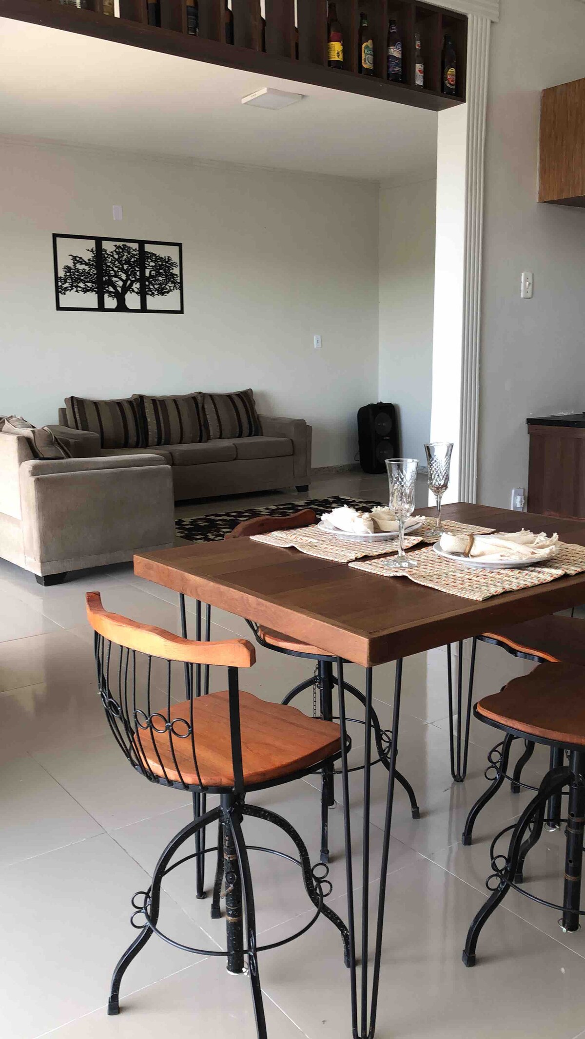 A dining area features a wooden table set with plates and glasses, complemented by four metal-framed chairs. In the background, a comfortable gray sofa with striped cushions is positioned against a wall adorned with a black and white tree print.