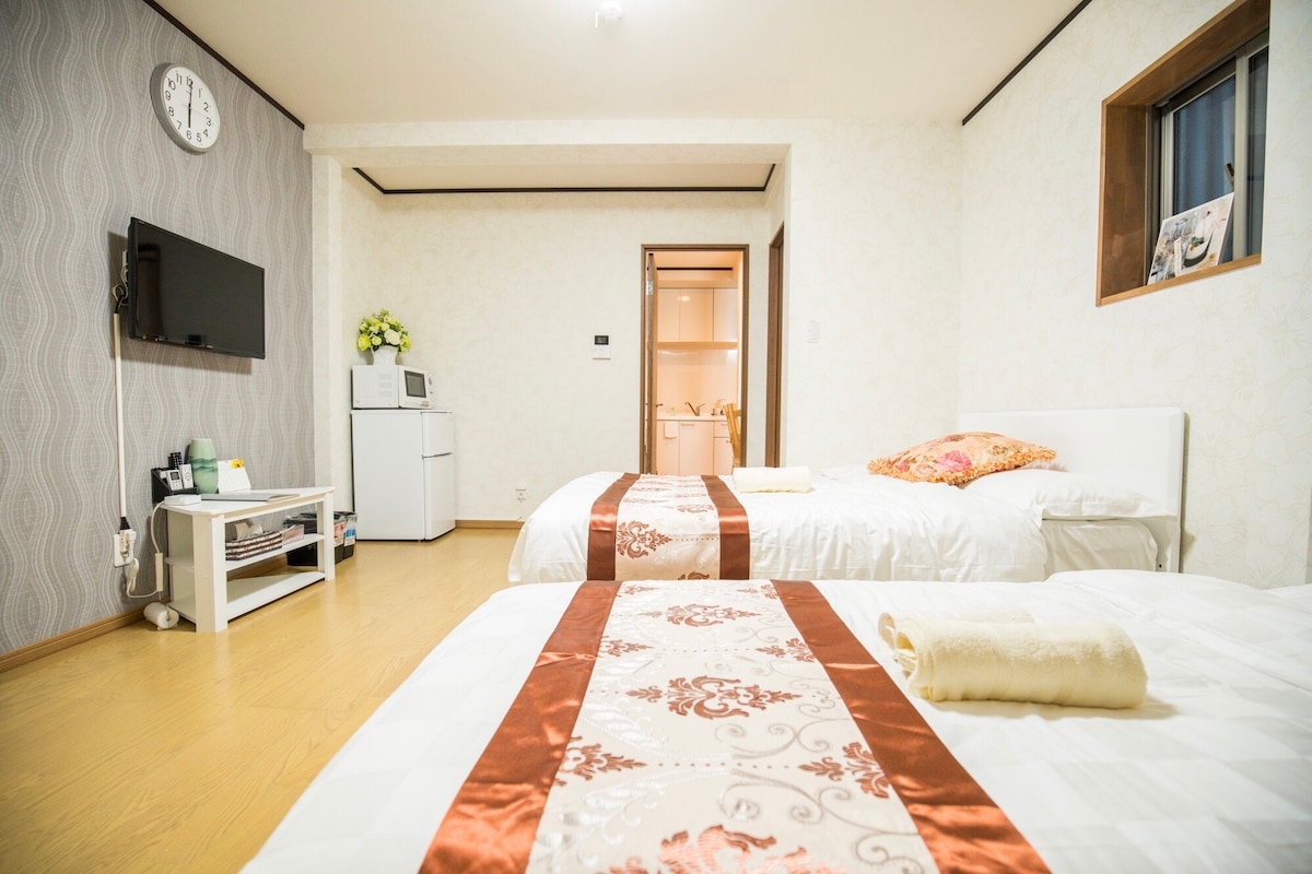 A well-lit sleeping area features two spacious beds adorned with patterned bed linens. A flat-screen TV is mounted on a light wall, beside a small dining table and chairs. A bright window offers natural light, with a glimpse of an adjoining bathroom area.