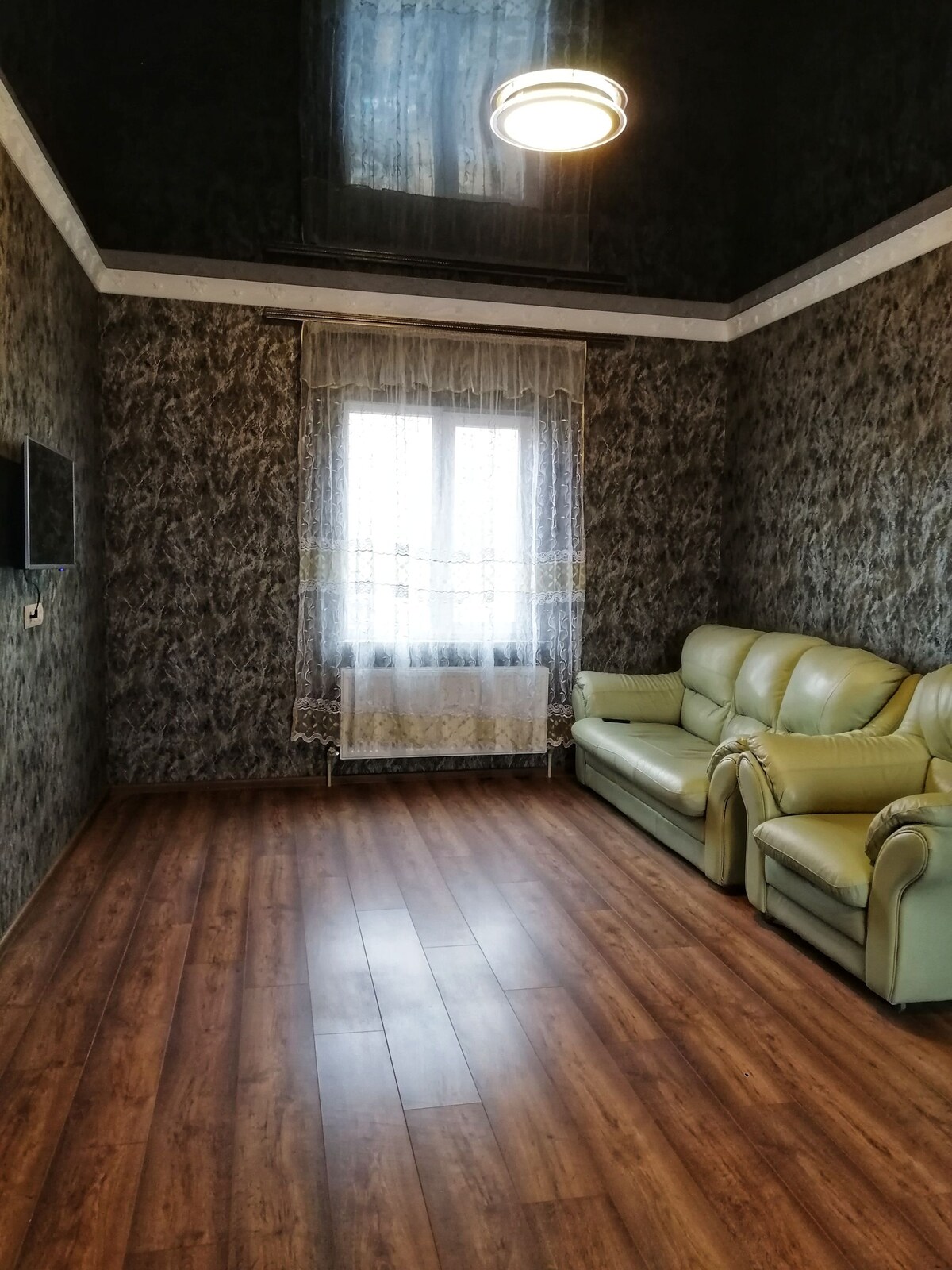 A sitting area is furnished with two light green reclining chairs facing a wall-mounted television. A large window, adorned with sheer lace curtains, allows natural light to fill the space, while textured dark wallpaper adds depth to the room.