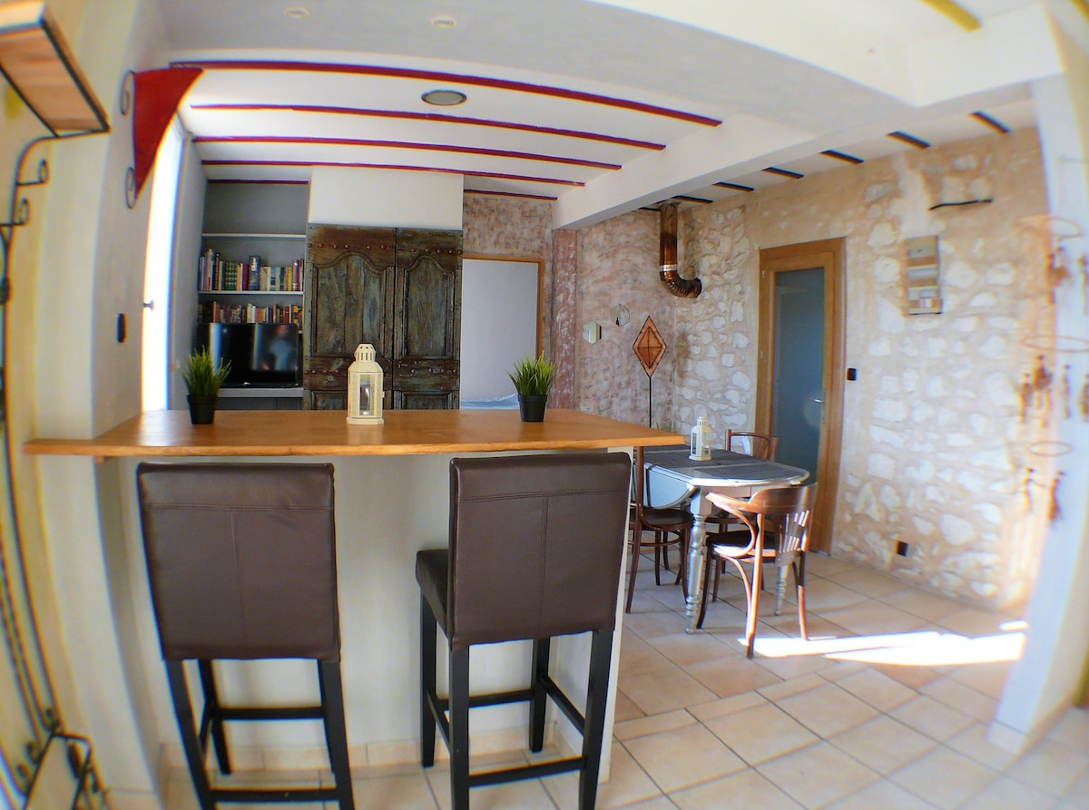 The interior features a bar area with two tall stools overlooking a small dining table and chairs. The wall highlights a mix of light stone and painted surfaces. A vintage cabinet can be seen in the background, complemented by shelves lined with books and decorations.