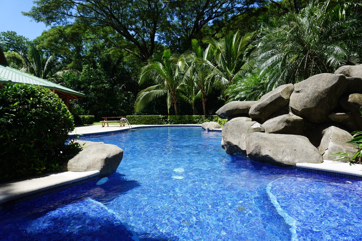 A tranquil pool is surrounded by lush tropical greenery and large rocks, creating a natural feel. The water is a vibrant blue, reflecting the sunlight filtering through the palm trees. A shaded area with seating can be seen in the background.