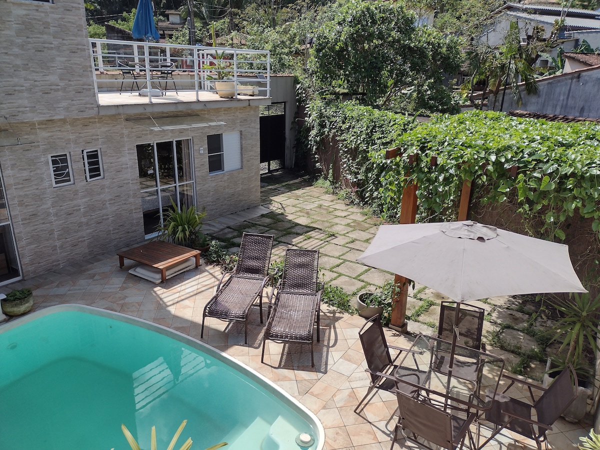 The outdoor area includes a refreshing pool surrounded by lounge chairs and dining furniture under an umbrella. Lush greenery is visible in the background, with stone pathways leading to a patio area. The setting promotes relaxation in a serene environment.