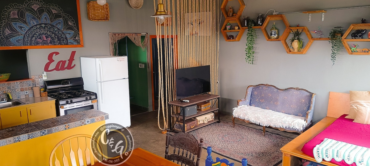 A vibrant living area showcases a mix of rustic and Boho design elements. A stylish sofa accompanies a wooden coffee table in front of a smart TV. The kitchen area features a gas stove and refrigerator, and hexagonal shelves display various decor items along the walls.