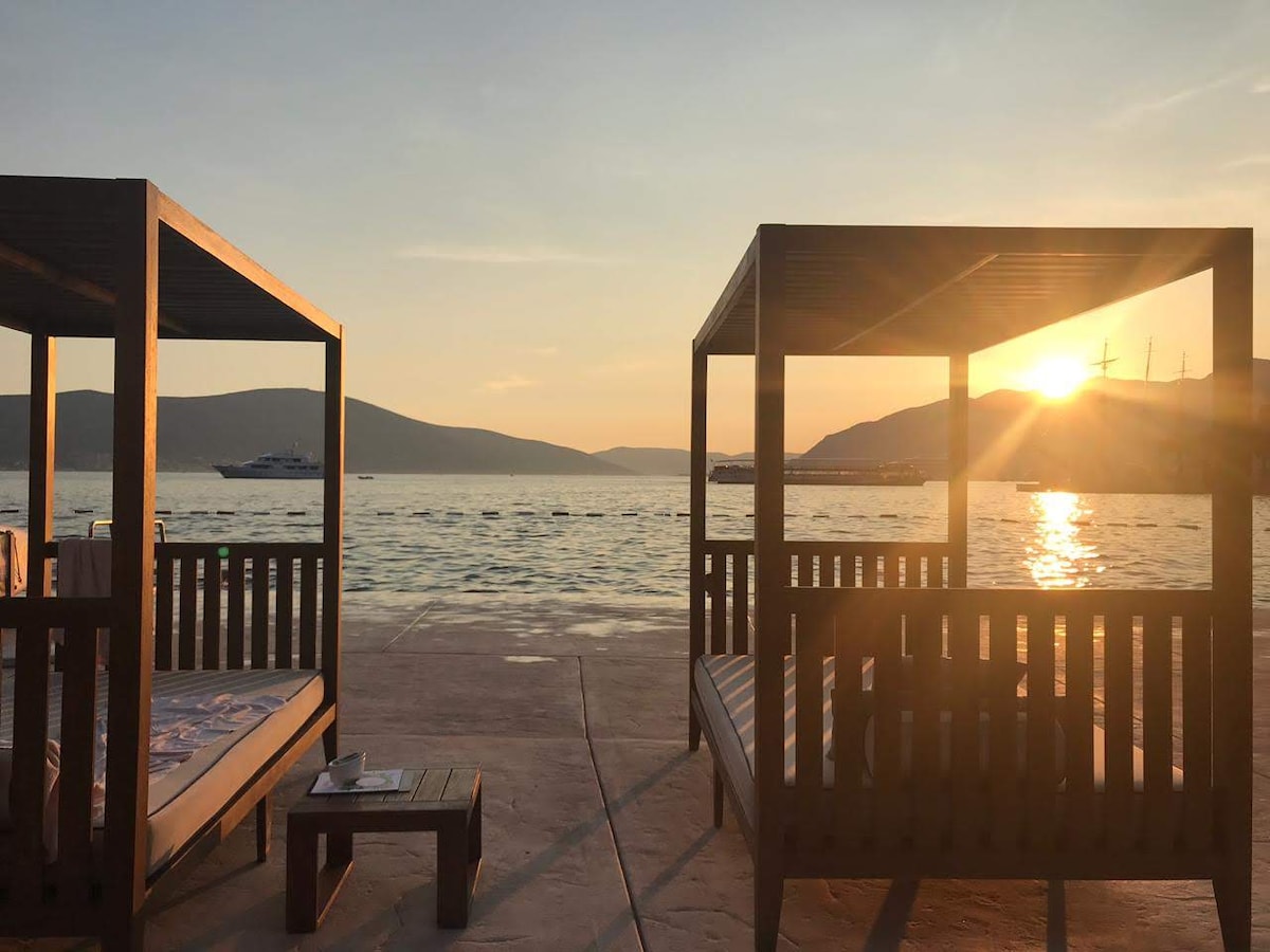 Two wooden cabanas are positioned on a waterfront terrace, framed by a serene sea view. The sun sets in the background, casting warm light over the water and mountains in the distance. A small table is placed beside the cabanas.