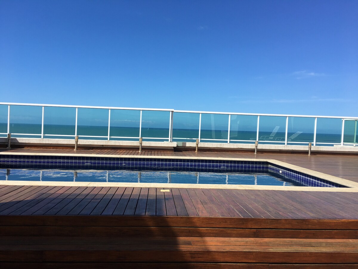 A serene outdoor area features a swimming pool surrounded by wooden decking. Glass railings provide an unobstructed view of the ocean and clear blue sky. Lounge chairs are positioned nearby, inviting relaxation in the sun.