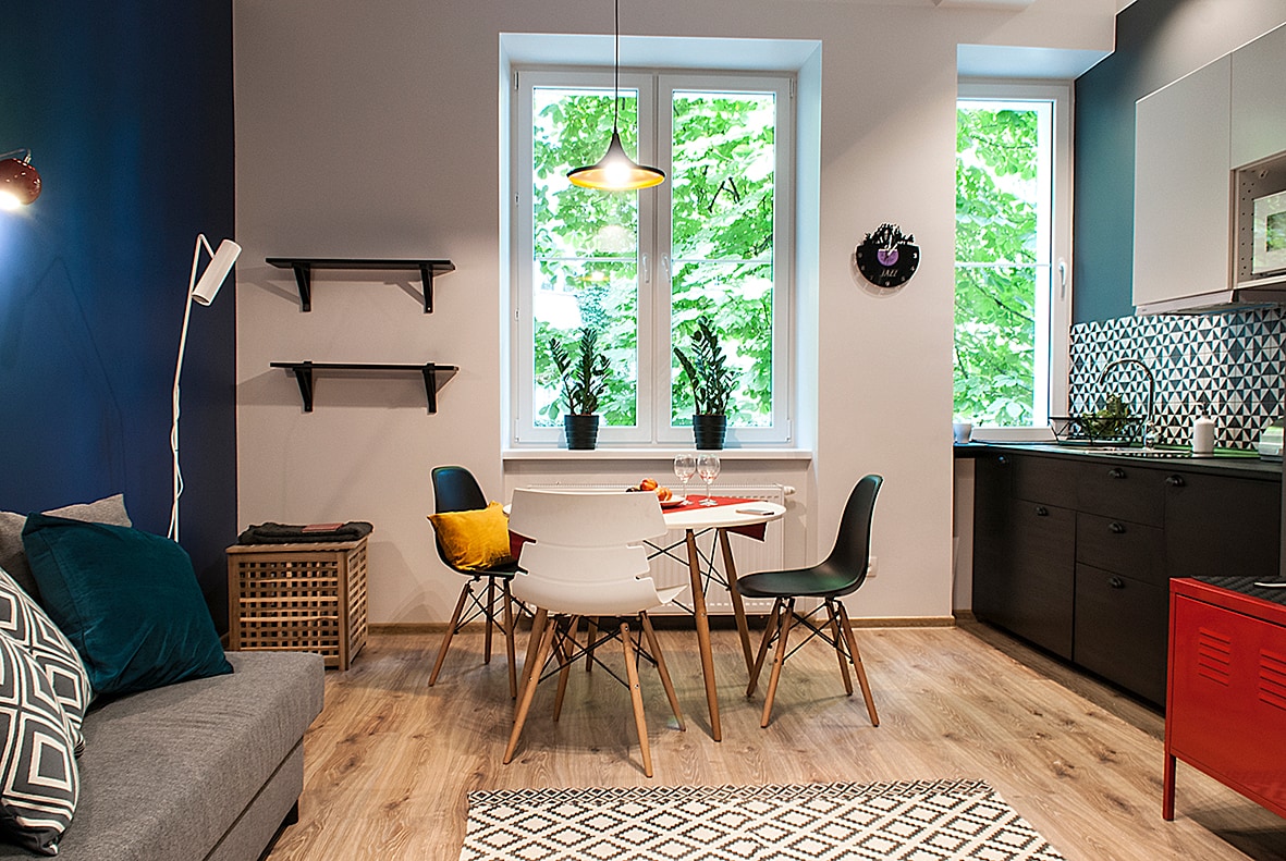 A cozy living area is featured, with a gray sofa adorned with colorful pillows. A round dining table surrounded by chairs is positioned near large windows, offering views of greenery. A well-equipped kitchen with dark cabinetry is visible in the background.