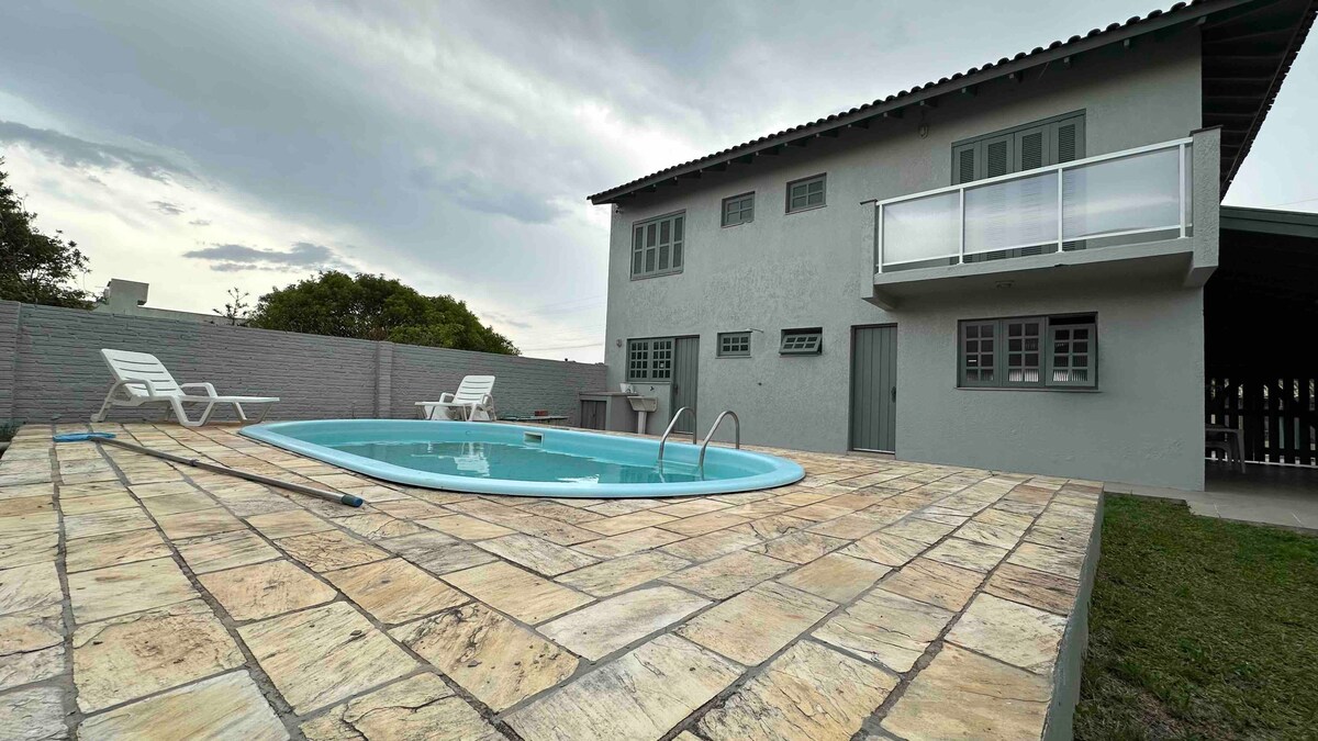 A spacious outdoor area features a swimming pool surrounded by a stone patio and white lounge chairs. The two-story house is visible in the background, with large windows and a balcony that enhances the overall design. Lush greenery is present around the perimeter.