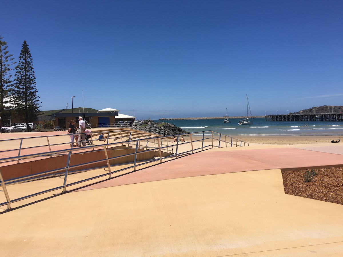 A gentle sloping pathway leads to a sandy beach, where sailboats rest in the tranquil water. Nearby, a lively recreational area is visible with shaded seating. Palm trees line the background, complemented by a clear blue sky, enhancing the inviting seaside atmosphere.