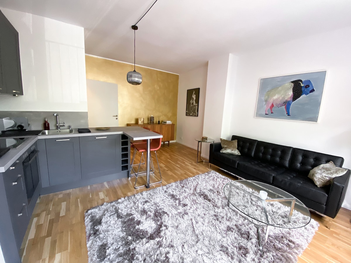 The open-plan living area features a modern kitchen with sleek cabinetry and a breakfast bar, complemented by stylish seating arrangements. A cozy gray rug enhances the space, while an eye-catching artwork adorns the wall, creating a contemporary ambiance.