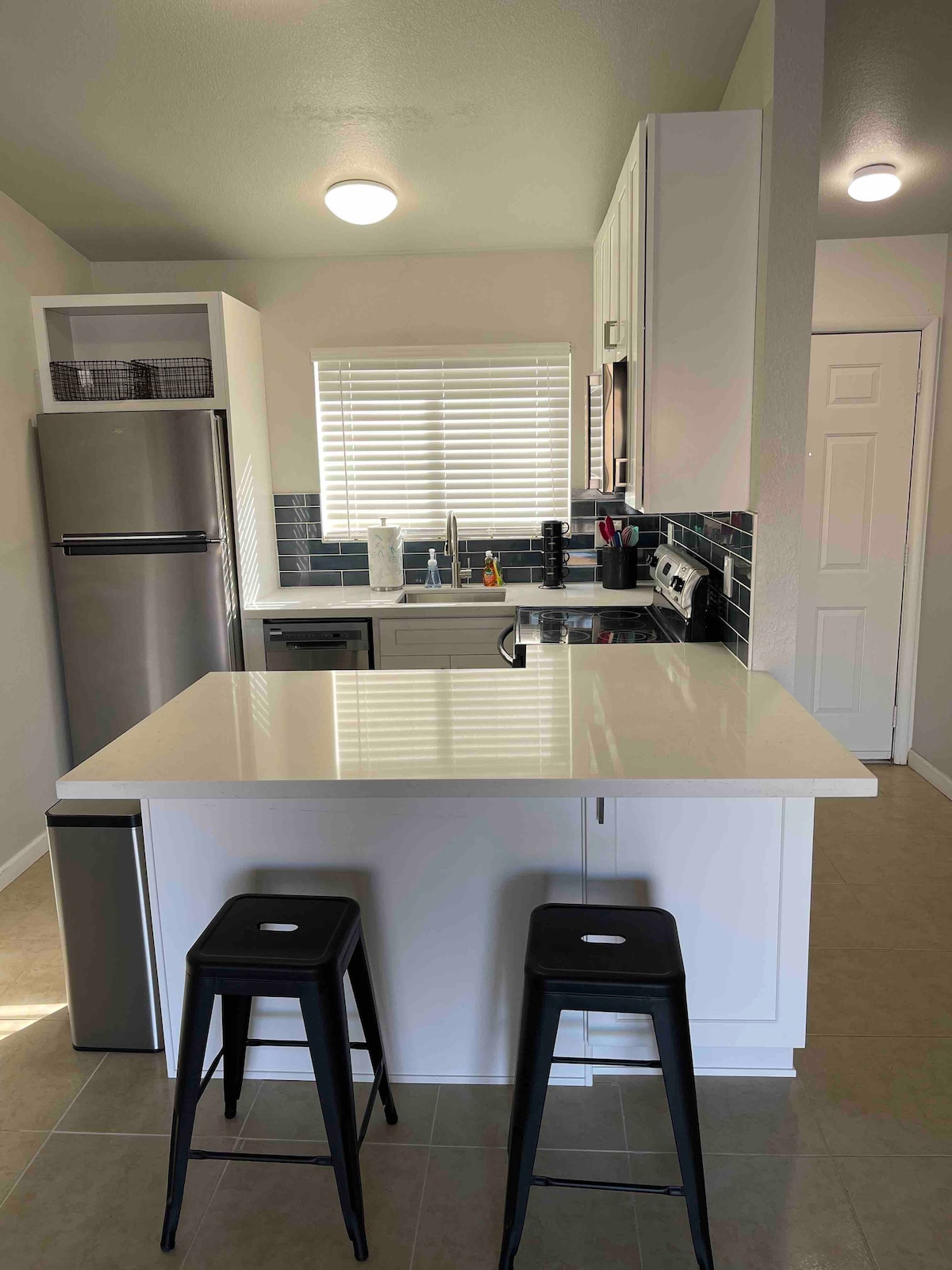 A modern kitchen features sleek cabinetry and stainless steel appliances. A central island with two black stools provides a casual dining option. Natural light from a window brightens the space, which is accented with dark backsplash tiles, creating a contrast with the light countertops.