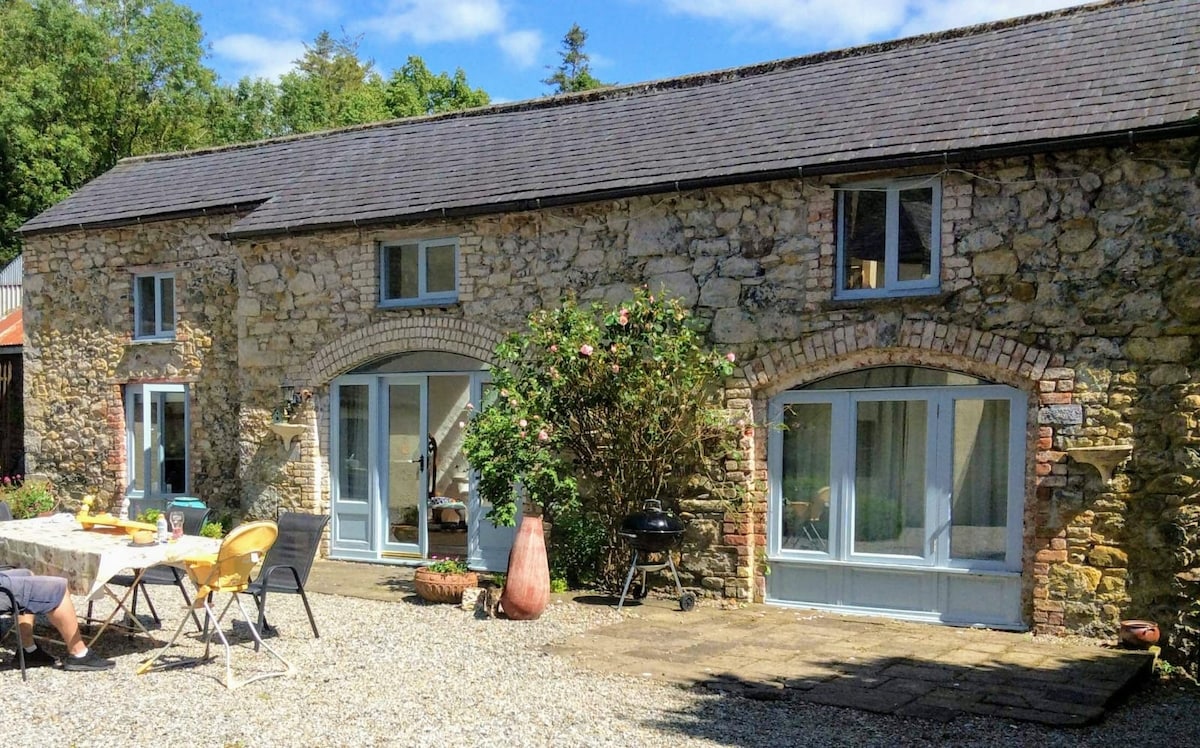 The exterior of the coachhouse is featured, showcasing a charming stone structure with large, light-filled windows. A spacious patio area includes outdoor seating and a charcoal barbecue, surrounded by gravel and greenery, creating a peaceful outdoor setting.