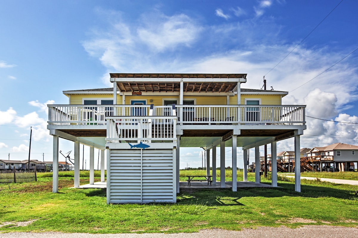 The elevated house features a spacious front deck with white railings, providing a shaded area beneath the roofline. Grass surrounds the structure, and the sky is partly cloudy, creating a bright backdrop. The design includes an open space underneath, ideal for outdoor activities.