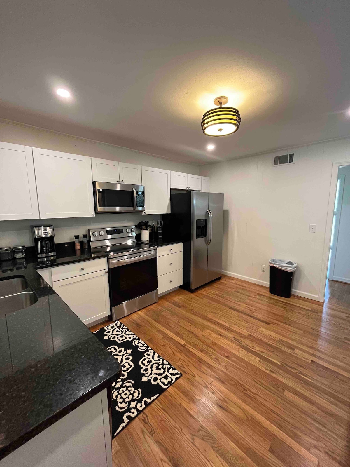 A modern kitchen features sleek white cabinetry with dark granite countertops. Stainless steel appliances, including an oven and a refrigerator, complement the design. The space is illuminated by a stylish ceiling light, while light wood flooring adds warmth to the environment.