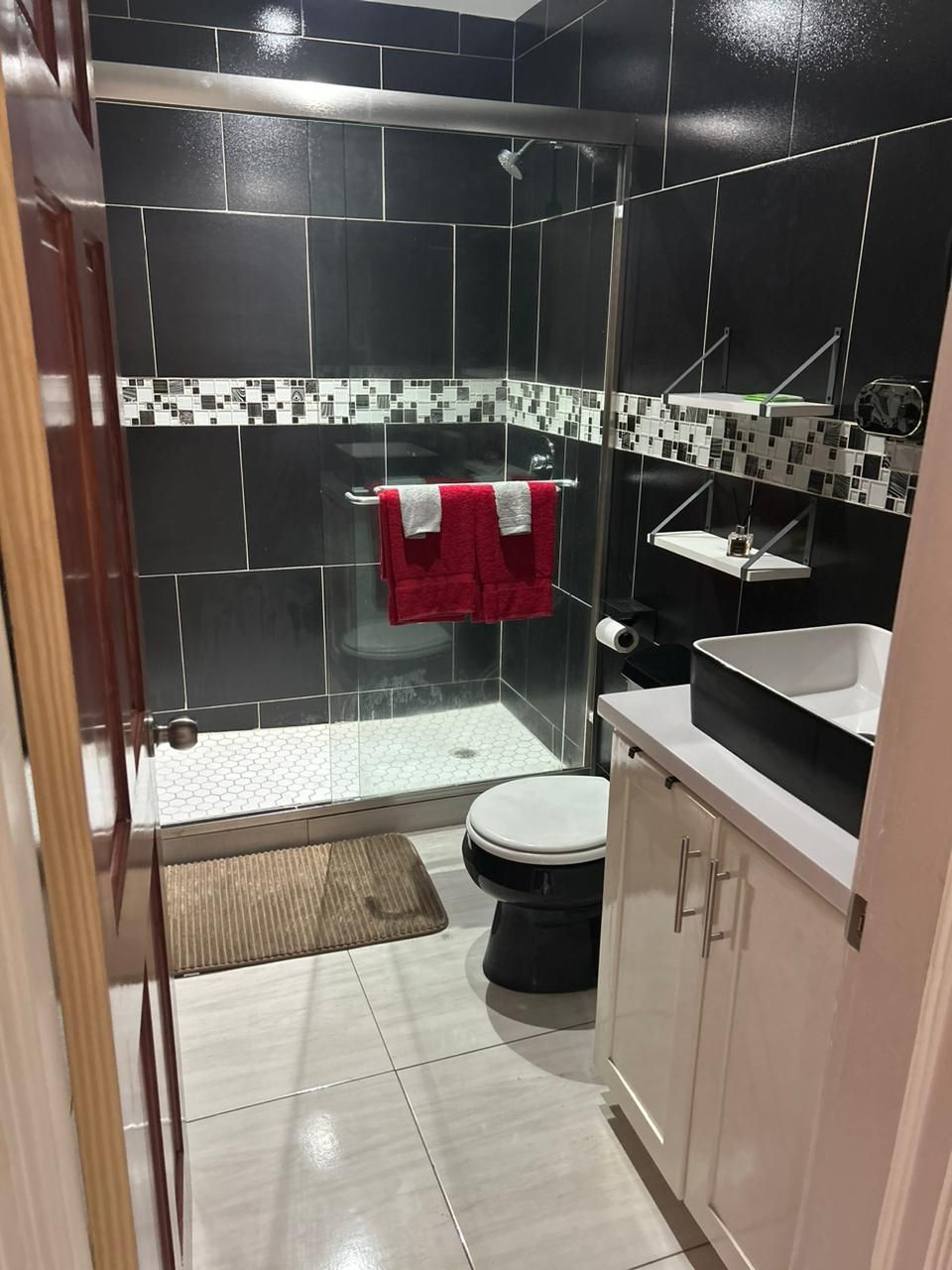 A modern bathroom features sleek black and white tiles, with a walk-in shower enclosed by clear glass. Fresh red towels are neatly arranged on a towel rack. Below the sink, a white cabinet offers storage, while a small mat is placed beneath the shower area.