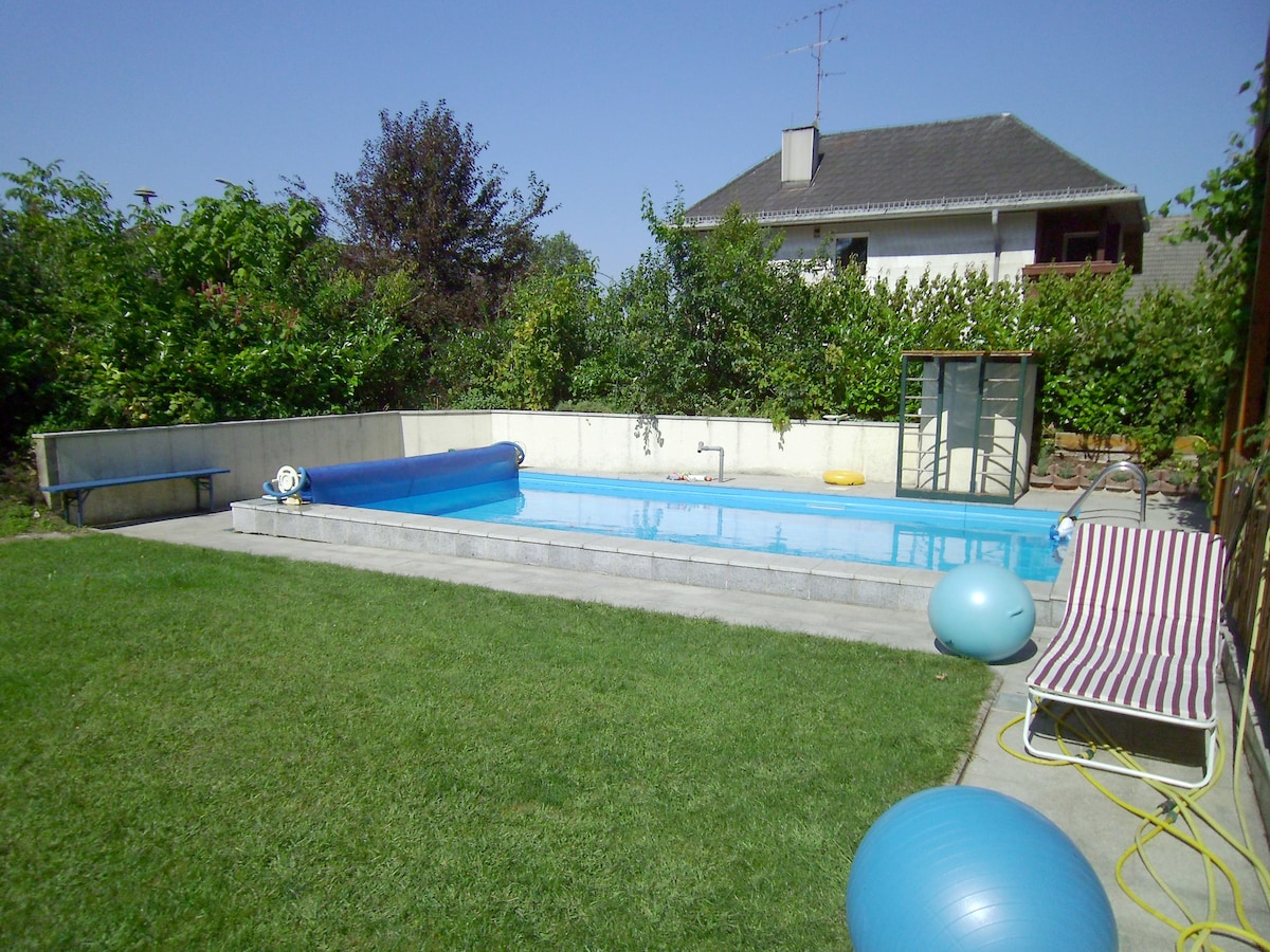 An outdoor pool is surrounded by a well-maintained green lawn. A covered patio area and shrubbery provide privacy, while colorful exercise balls are positioned nearby. A striped lounge chair offers a spot to relax, and a retractable cover is rolled up at one end of the pool.
