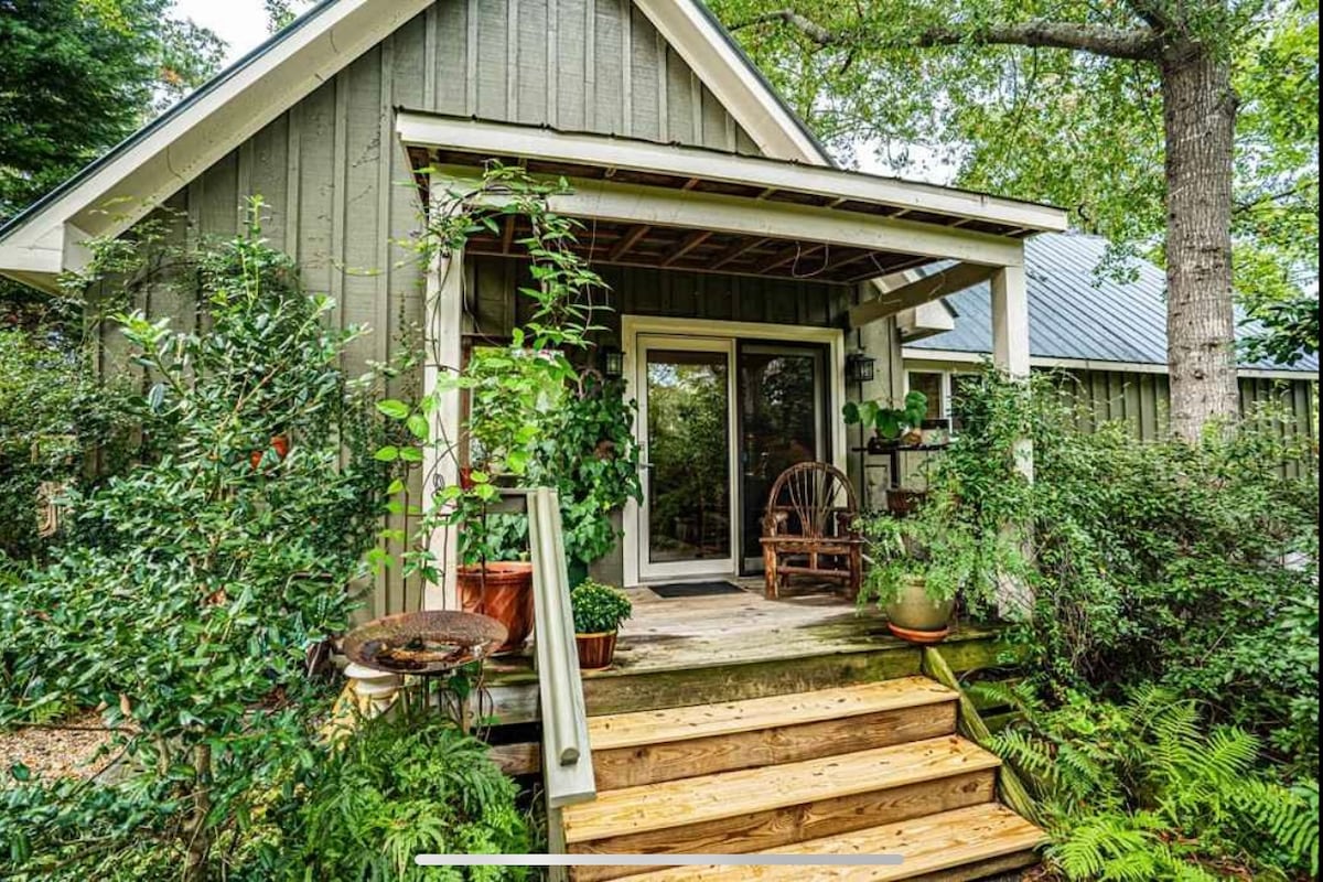The entrance features a welcoming porch with wooden steps leading to a glass door. Potted plants and thriving greenery surround the porch, offering a serene atmosphere. Rocking chairs provide a comfortable spot to enjoy the natural beauty of the surroundings.