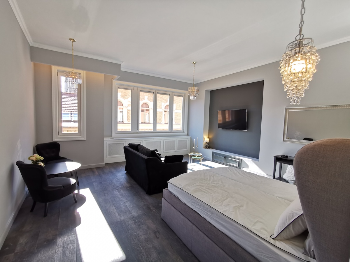 A spacious room features a large bed and a cozy seating area with a black sofa and armchairs. Natural light enters through large windows, enhancing the light-colored walls. Decorative elements include modern light fixtures and a wall-mounted television.