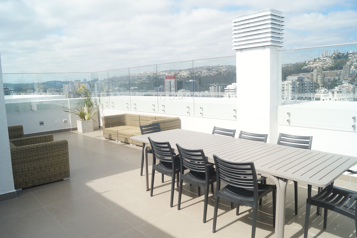 An outdoor terrace is presented with a long dining table surrounded by black chairs. Comfortable lounge seating is arranged nearby. The area is framed by glass panels, offering panoramic views of the city and mountains in the distance under a partly cloudy sky.